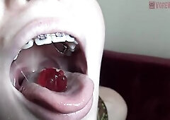 Blue- eyed golden-haired with braces is licking and eating gummy bears in a very hot way